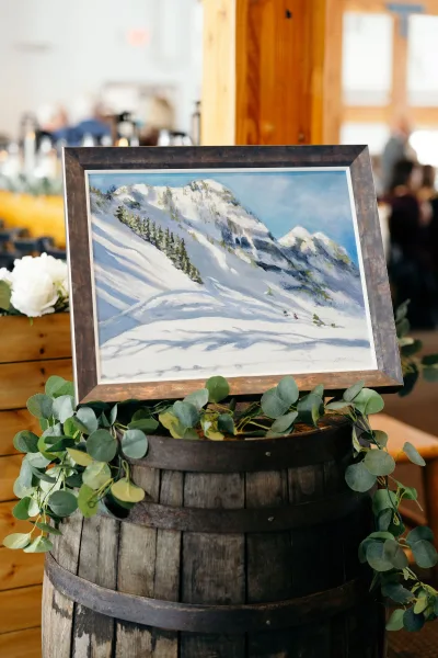 Wedding welcome table decor on a wooden barrel with framed artwork and eucalyptus garland, set in a warm wooden interior with guests blurred behind