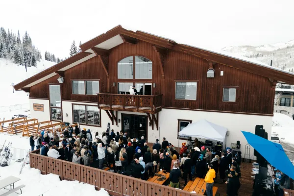 Outdoor wedding ceremony on a ski lodge deck, guests in winter coats beneath a canopy tent with snowy mountains behind