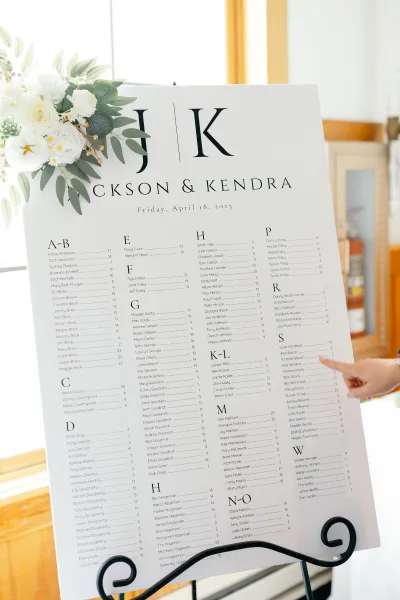 Seating chart wedding seating chart sign on a black metal easel, accented with white florals and greenery in window-lit venue