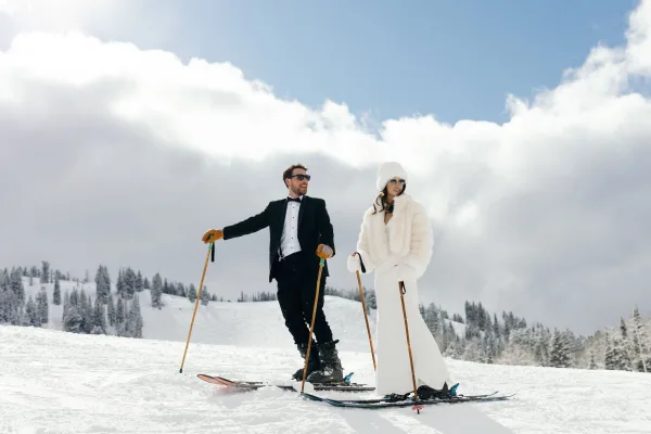 Couple portrait in ski wedding photos, bride in fur coat and groom in tuxedo on skis with pine trees and snowy mountains behind