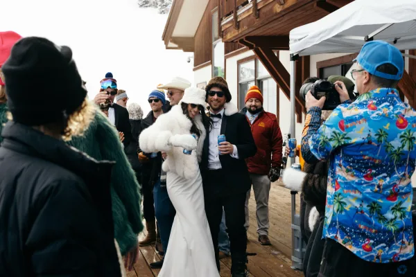 Couple portrait of bride in wedding dress and faux fur jacket with groom in tuxedo and sunglasses on a lodge deck by a canopy crowd