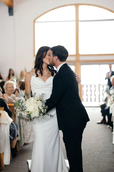 Wedding kiss as bride in white dress and groom in black tuxedo kiss in an indoor chapel aisle, guests seated by a large arched window