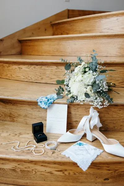 Wedding details flat lay with wedding invitation suite, rings in a box, pearl jewelry, white heels, bouquet, on a wooden staircase