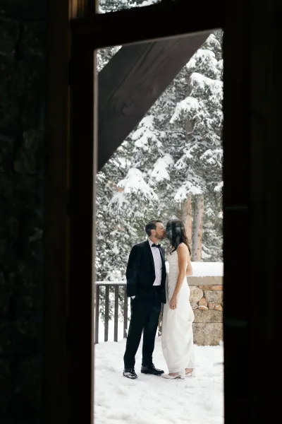 Wedding kiss portrait of bride and groom kissing, her strapless dress and heels beside his tuxedo on a snowy cabin porch with evergreens