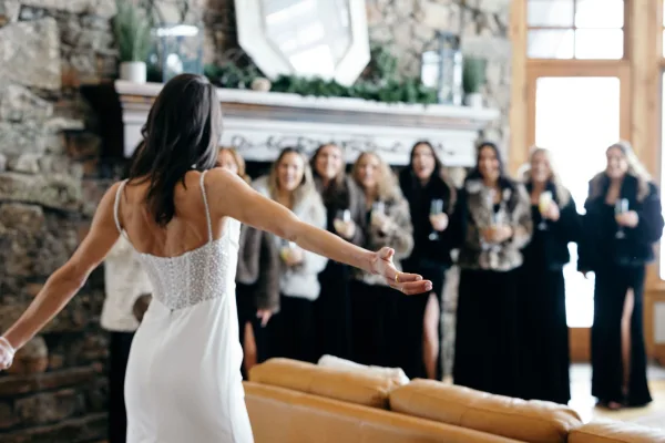 Bridal party toast as the bride in a beaded wedding dress cheers with bridesmaids holding champagne flutes by a stone fireplace in a living room