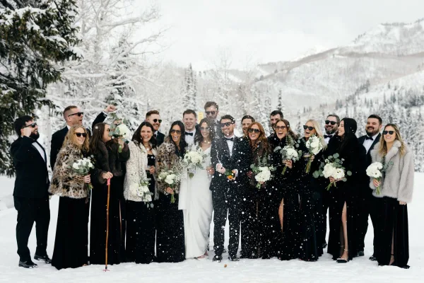 Wedding party photo in snow with bride and groom popping champagne, sunglasses and tuxedos amid snowy trees and winter mountains