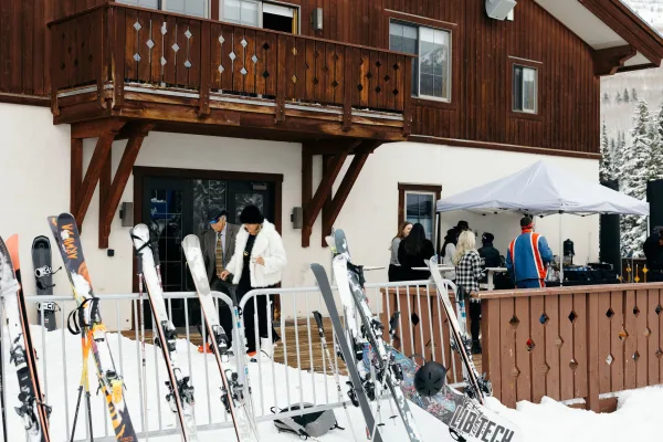 Après ski wedding guests mingle in winter jackets beside skis, sipping drinks at cocktail tables outside a wood chalet on snowy slopes