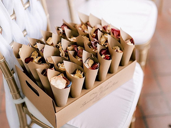 Petal cones for wedding petal toss filled with dried rose petals in paper cones on a cardboard tray beside a ribboned chair