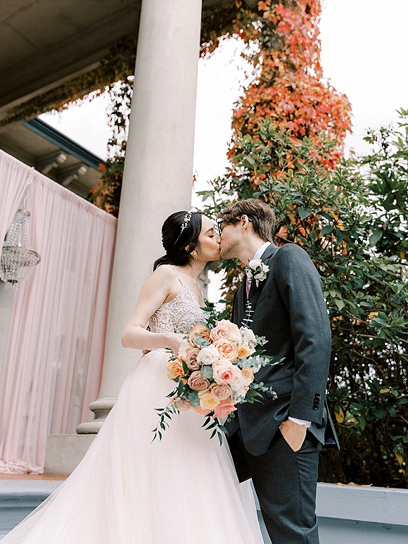 Wedding kiss as the bride and groom kissing on outdoor steps, her blush bouquet and beaded headband framed by columns and ivy