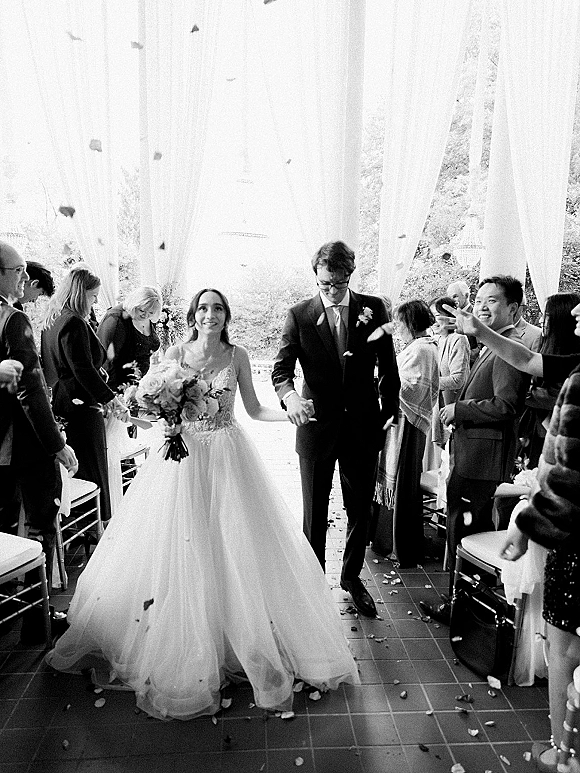 Wedding recessional as newlyweds walking down aisle holding hands, bride with bouquet while guests cheer and toss flower petals indoors