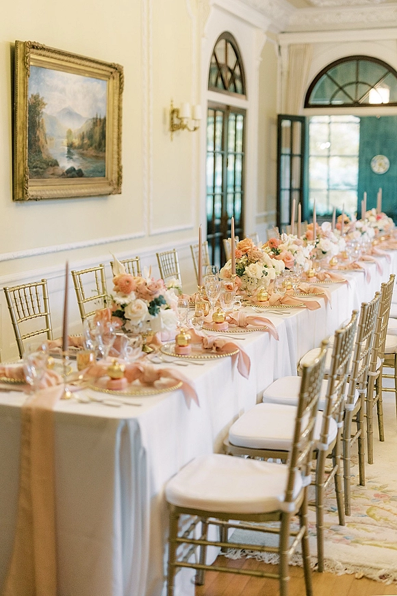 Reception tablescape with long head table decor, blush napkins, floral centerpieces, and taper candles in an elegant white-paneled room