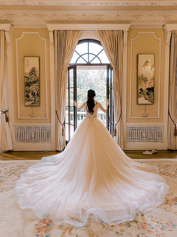Bridal portrait of a bride back view in a ball gown wedding dress with lace bodice and long train, standing by French doors in a classic room
