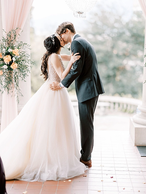 Wedding kiss portrait of bride and groom kissing in side profile, bride in lace bodice and tulle skirt, under chandelier on terrace columns
