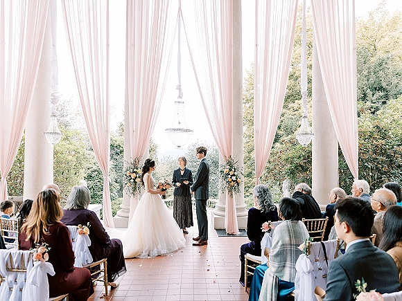 Wedding ceremony with couple exchanging vows beneath blush drapery and florals on a columned veranda, rose petal aisle and chandeliers overhead