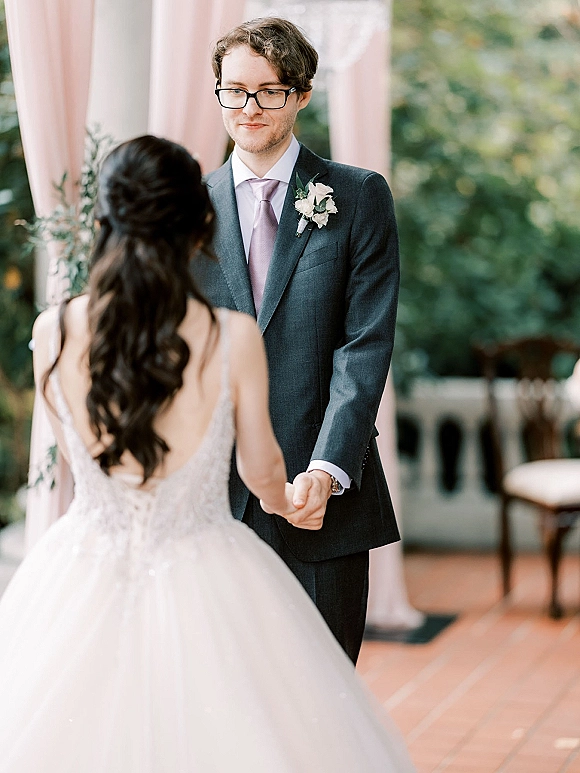 Wedding vows as the couple holding hands, groom in gray suit with glasses and lilac tie beside bride in backless lace gown outdoors with greenery and blush draping