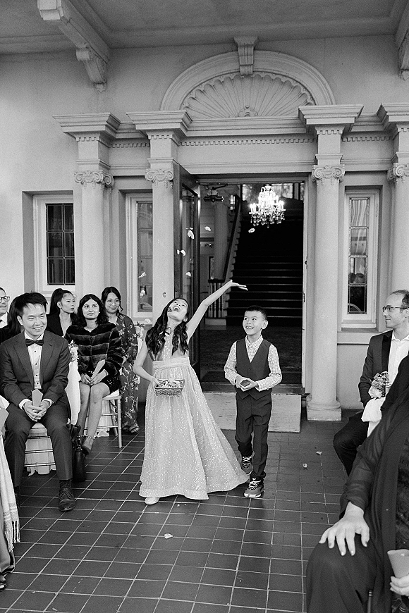 Flower girl moment as she tosses petals from a basket, walking past seated guests at a columned building entrance with chandelier