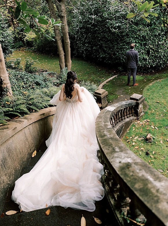 Wedding first look as bride approaching groom from behind, her ball gown with long train flowing down stone steps in a garden setting