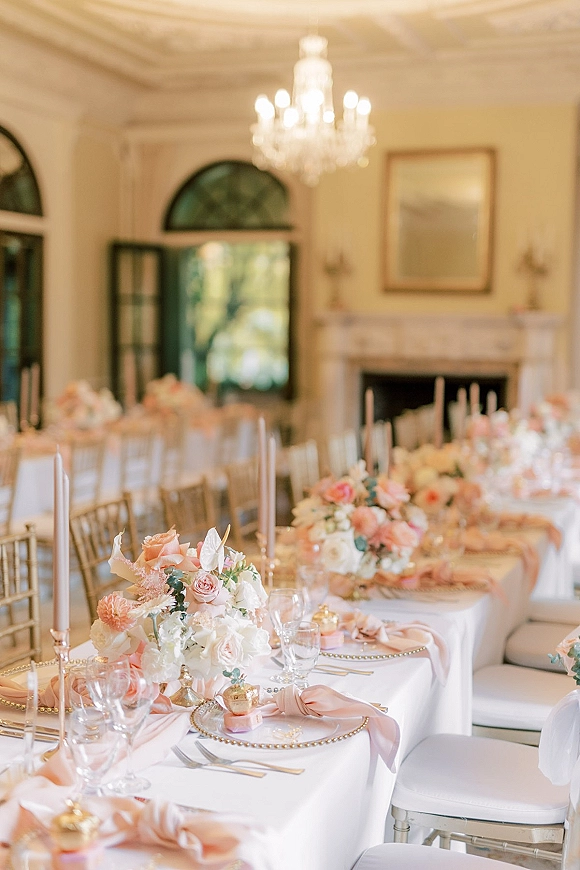 Reception tablescape with blush wedding tablescape details—white linens, pink florals, taper candles, gold chargers and chairs beneath a chandelier