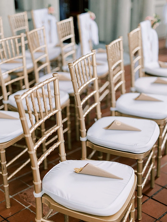 Ceremony seating with gold chiavari chairs and white cushions, paper confetti cones tied with ribbons on an outdoor brick patio aisle