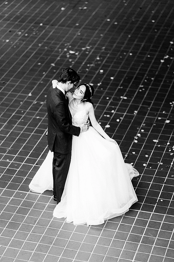 Couple portrait in a black and white wedding portrait, bride in tiara and veil embraced by groom in tuxedo on tiled floor with petals