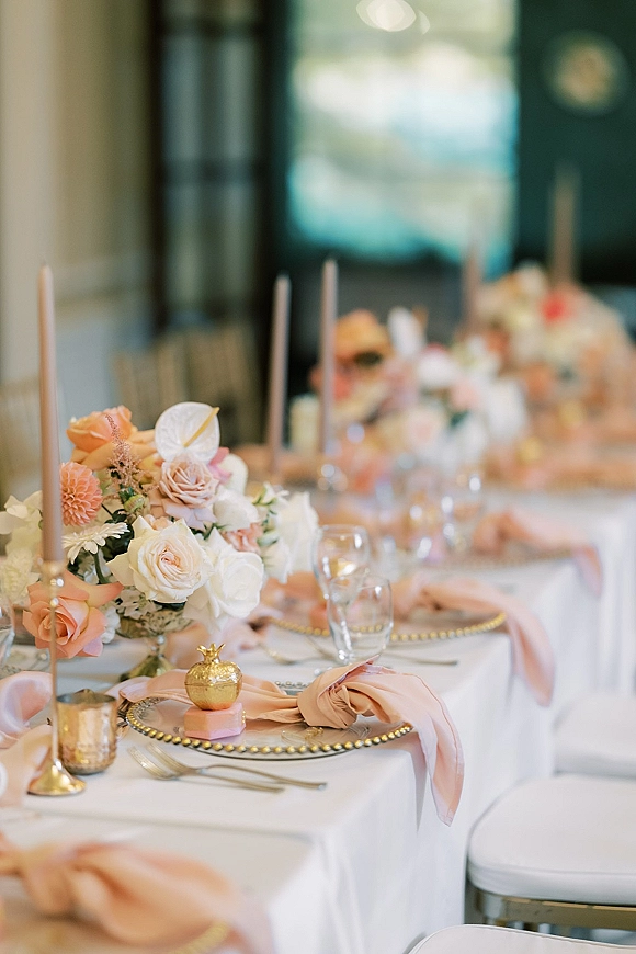 Reception tablescape wedding table setting with roses and dahlias, blush napkins, gold beaded chargers, and taper candles in an indoor space