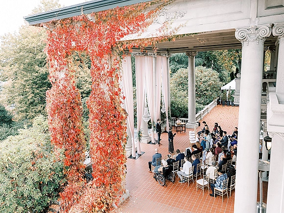 Outdoor wedding ceremony with white drapery and a chandelier, gold chiavari chairs on a columned veranda with autumn vines