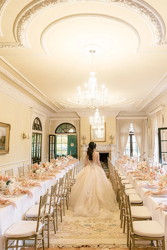 Reception tablescape with long banquet table setup, white linens, blush napkins, floral centerpieces, taper candles under crystal chandeliers