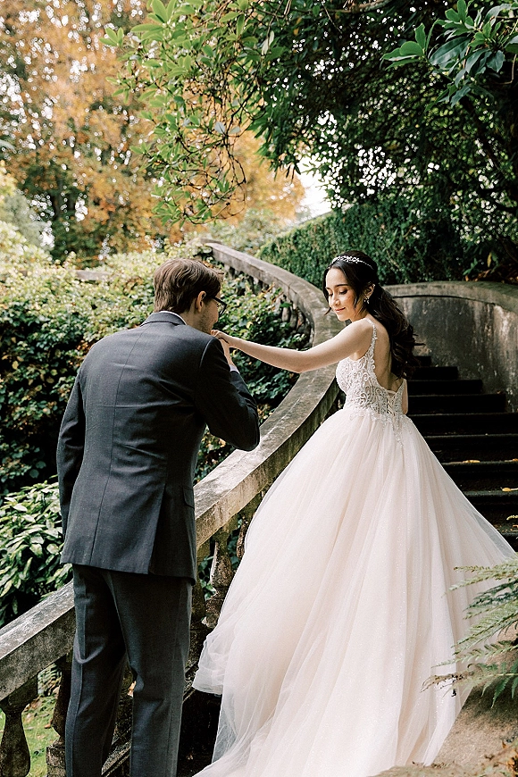 Couple portrait of groom kissing bride’s hand on a stone staircase, her ball gown and headband framed by lush garden foliage