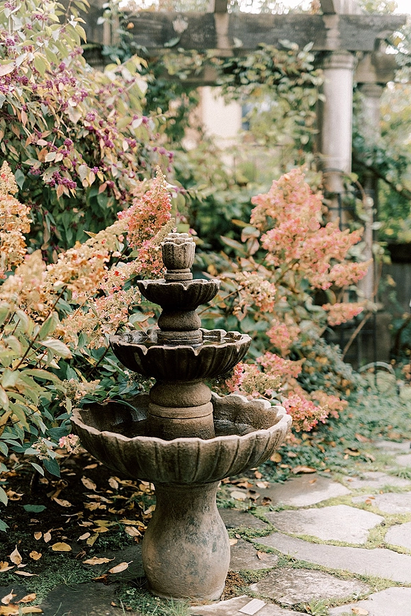 Garden fountain with a stone fountain centerpiece surrounded by flowering shrubs and vines near pergola columns on a stone path