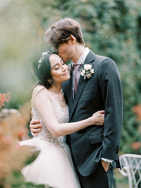 Couple portrait with a forehead kiss wedding moment as the groom embraces the bride in a beaded dress and hair vine, soft garden bokeh behind