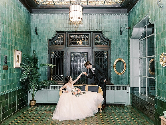 Couple portrait of bride in strapless ball gown on a sofa as groom kisses her hand, with bridal bouquet and stained glass backdrop