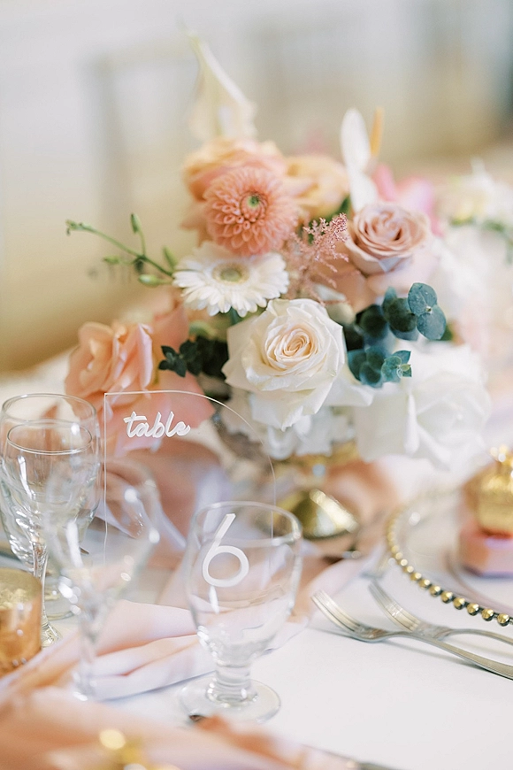 Reception tablescape with wedding table centerpiece of roses, dahlias and eucalyptus, acrylic table number, pink napkins, and votive candle on white linen