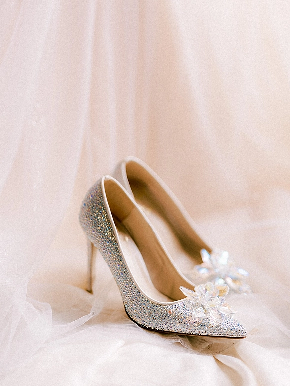 Bridal shoes with rhinestone wedding heels sparkle, featuring a crystal flower on pointed toes atop softly draped tulle fabric