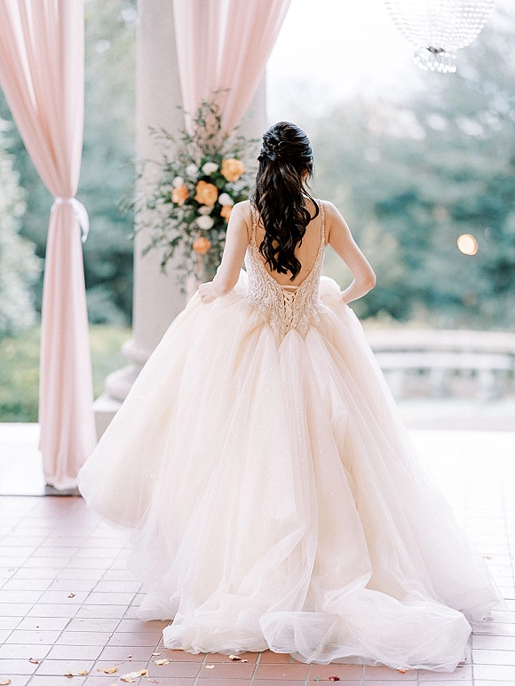 Bridal portrait of a bride in a backless wedding dress with lace bodice and long train, standing on a garden veranda with stone columns and chandelier.