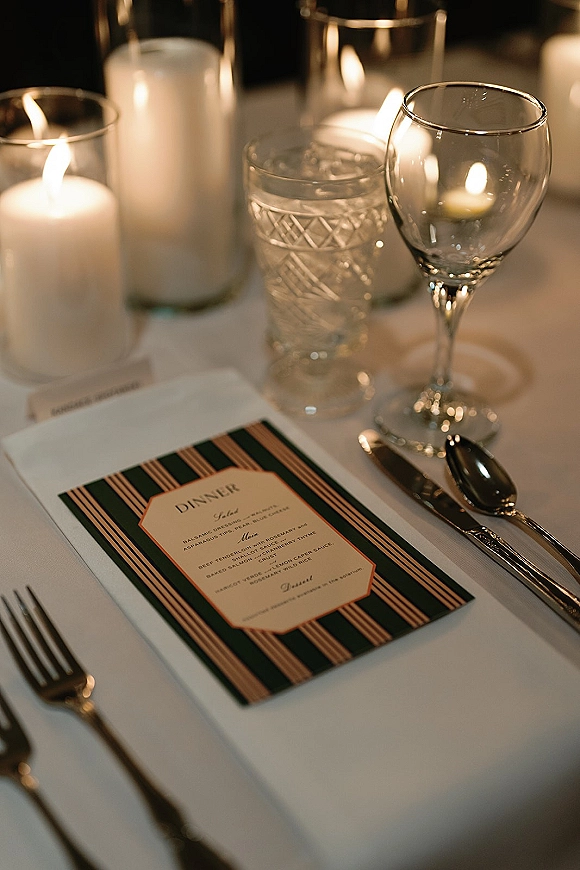 Reception tablescape with wedding place setting, striped black and gold menu card, silver flatware, wine and water glasses, and pillar candles in hurricanes