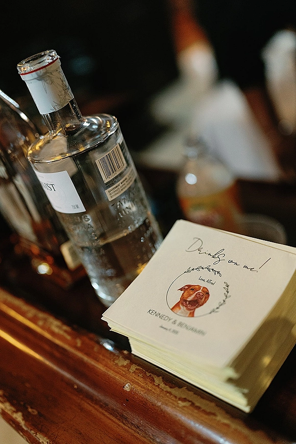 Wedding cocktail napkins, personalized cocktail napkins with calligraphy and a pet illustration, stacked on a wooden bar near liquor bottles