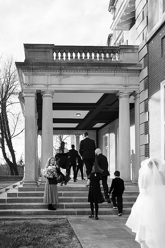 Wedding processional with bridesmaid holding bouquet as the wedding party walks up stone steps to a columned brick building, veil flowing