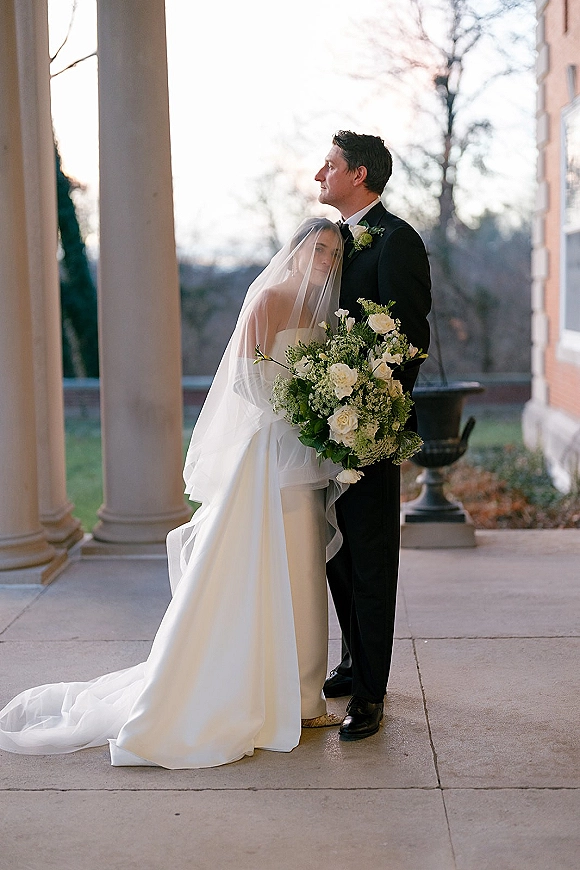 Couple portrait of bride leaning on groom, holding white rose bouquet with greenery, on a stone porch with columns and brick backdrop