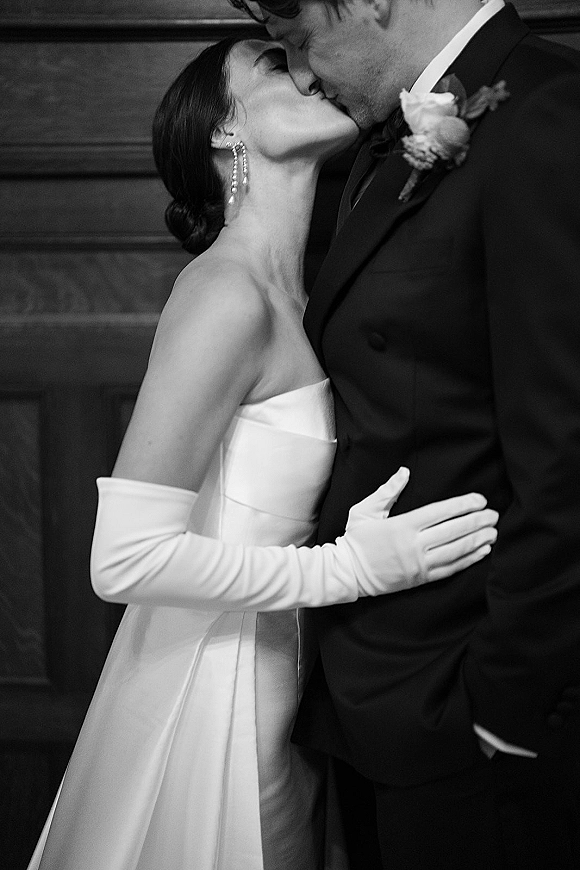 Wedding kiss portrait in black and white of bride in strapless dress and opera gloves kissing groom in glasses by wood-paneled wall