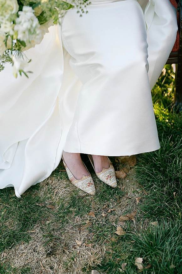 Bridal shoes wedding flats with floral embroidery under an ivory dress hem, beside white bouquet greenery on grass with fallen leaves