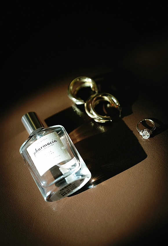 Wedding rings with engagement ring close up beside a perfume bottle and black ribbon on brown leather, lit by soft sunlight shadows