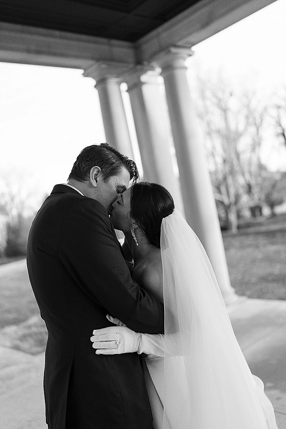 Wedding kiss portrait of bride and groom kissing, her veil and strapless dress visible, embracing beneath porch columns and trees