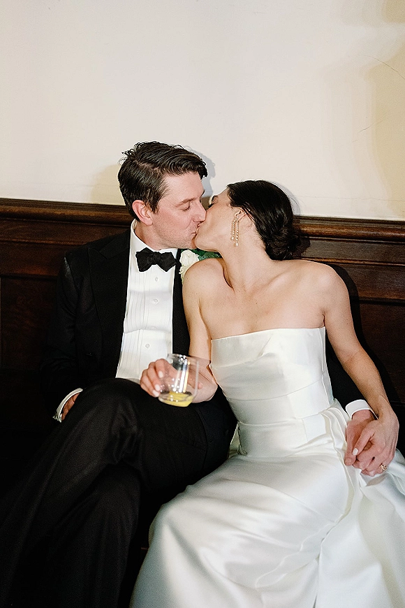 Wedding kiss portrait of bride and groom kissing on a wood bench, her strapless satin dress beside his black tuxedo against a cream wall