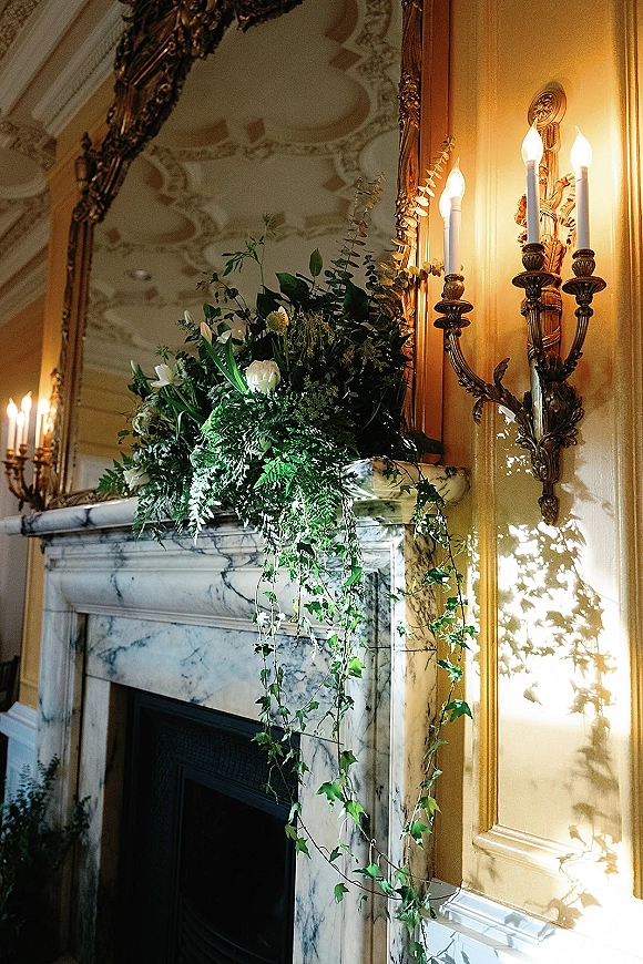Wedding mantel decor with fireplace mantel flowers, greenery garland and trailing ivy, white blooms, candles, and ornate mirror above marble fireplace