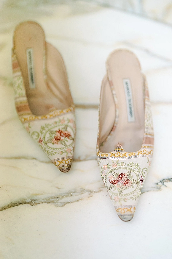 Bridal shoes with floral embroidered mules and pointed toes arranged on a marble surface, showcasing delicate pastel stitching