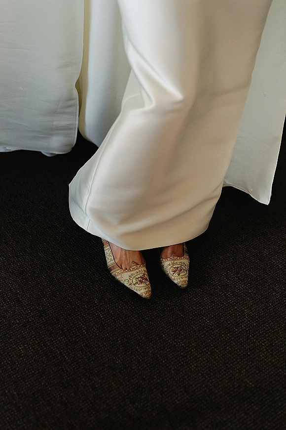 Bridal shoes in a wedding shoes close up, pointed-toe embroidered heels beside the bridal gown hem on white bedding and carpet