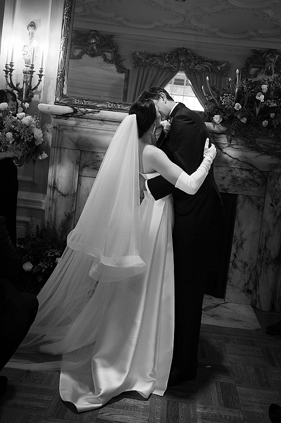 Wedding kiss portrait of bride and groom kissing, her veil trailing, in a black-and-white indoor room by a marble fireplace and mirror
