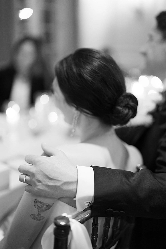 Reception moment of a wedding couple at reception, groom arm around bride, her low bun and drop earrings by candlelit tables and guests
