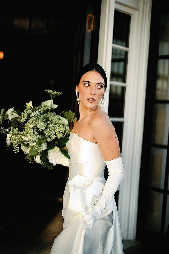 Bridal portrait of a bride in a strapless wedding dress with opera gloves, holding a white and green bouquet in a doorway with dark interior