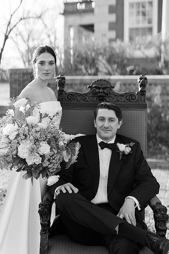Couple portrait in a black and white wedding portrait, bride in strapless gown standing behind groom seated on an ornate chair on a terrace